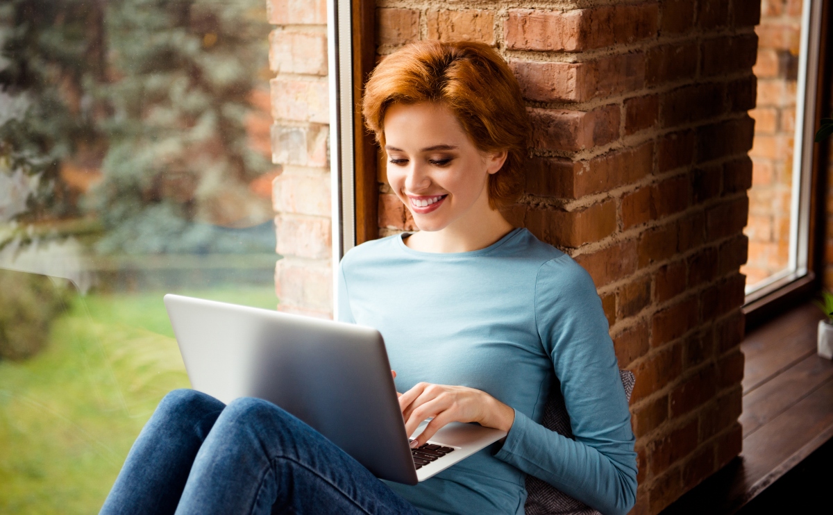 Pretty woman with short red hair sitting by a large window while checking her laptop