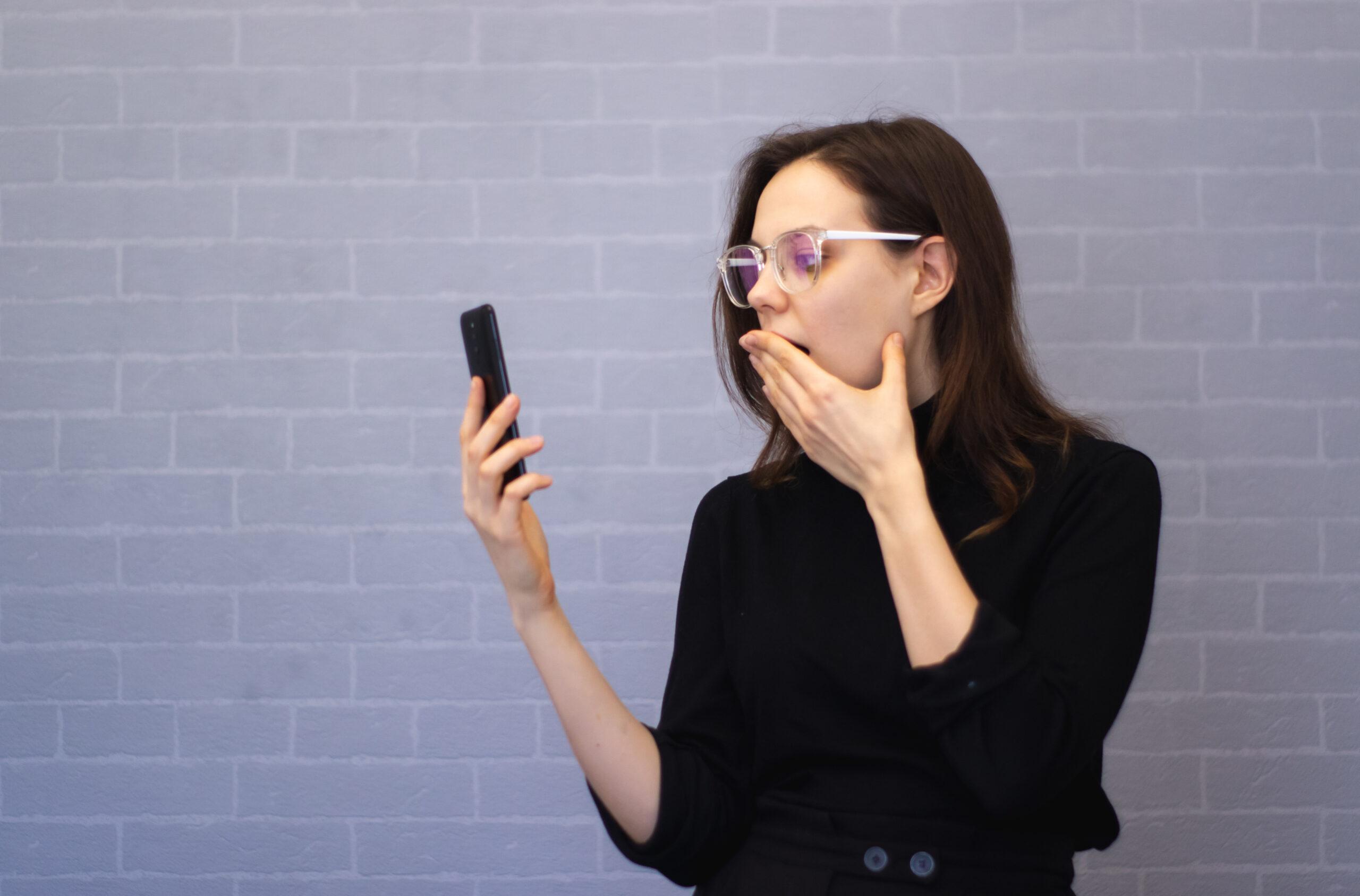 Shocked young woman using mobile phone cover mouth with hand 