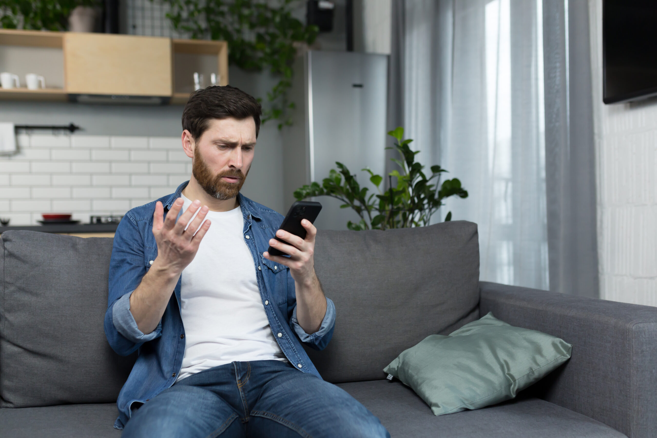 Thinking man looking at phone screen sitting on sofa at home