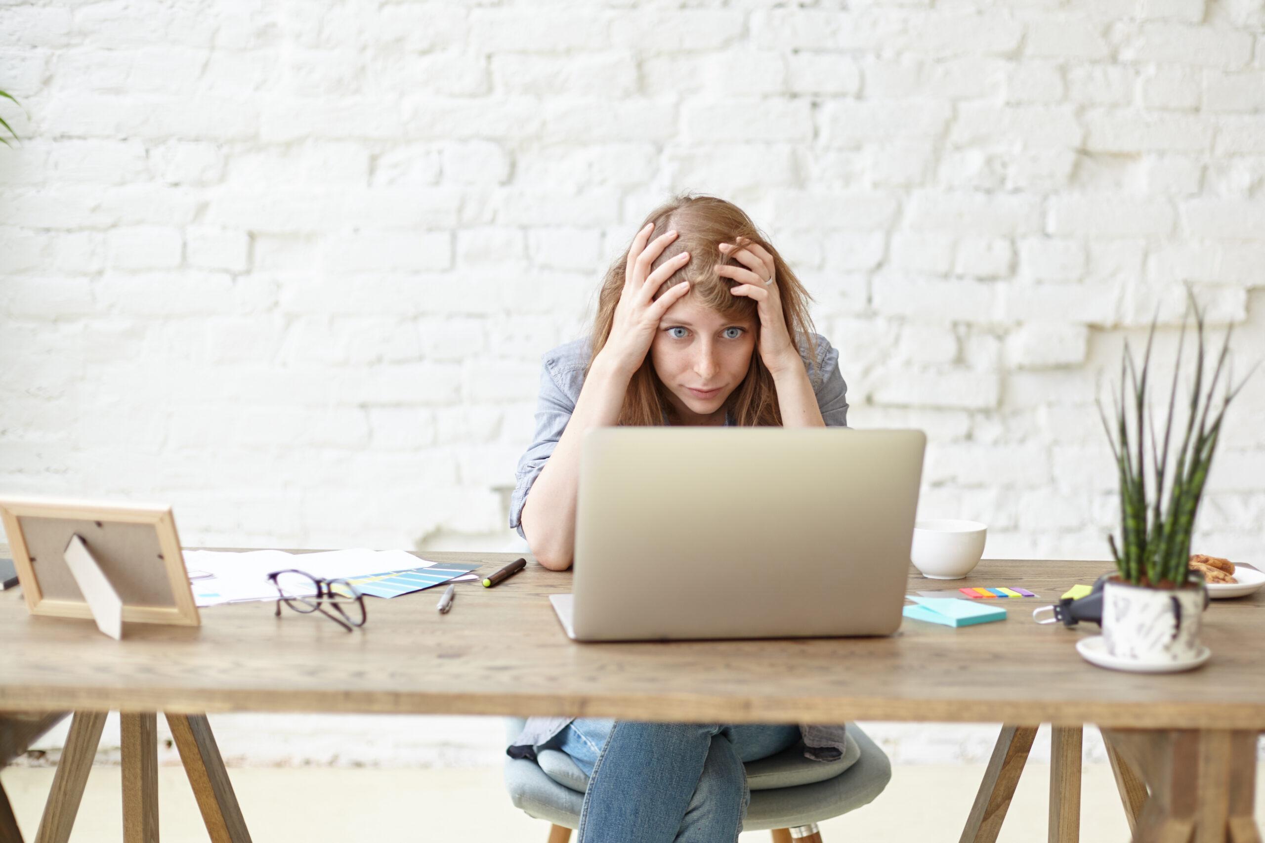 Indoor shot of frustrated female graduate student in casual clothes keeping hands on head, feeling panic and stressed out while working on diploma project, facing deadline, using laptop computer