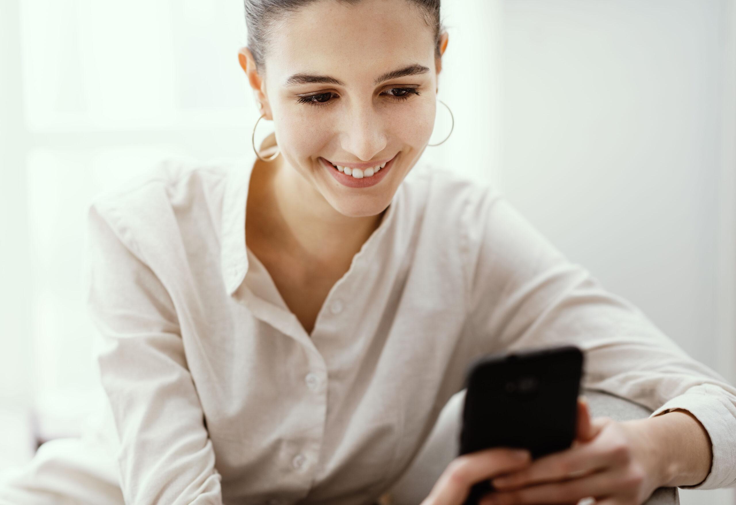 Happy young woman relaxing at home she is sitting on the armchair and chatting with her touch screen smartphone