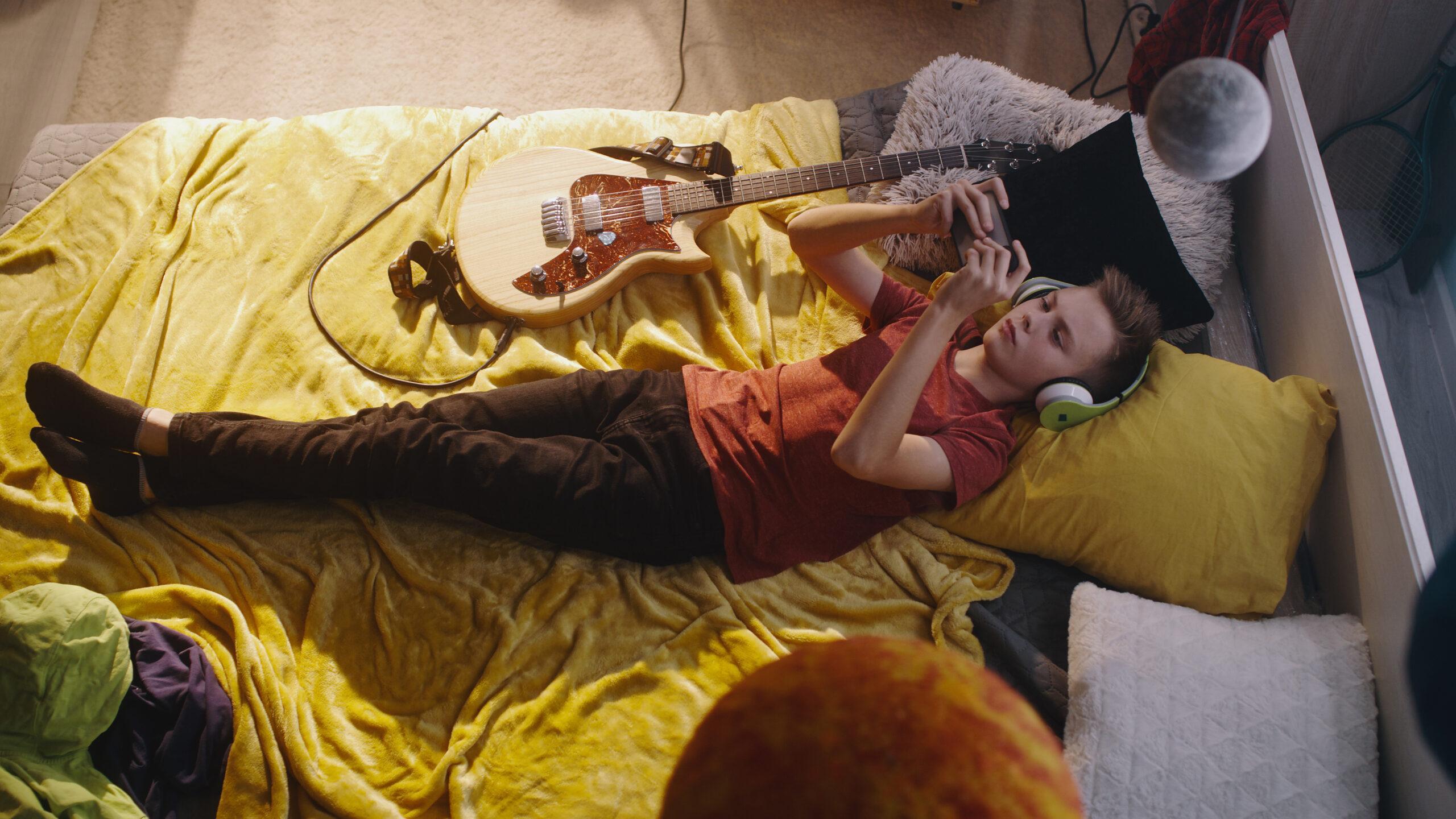Teenager in casual clothes and wireless headphones lying on bed near electric guitar and playing videogame on mobile phone in bedroom at home