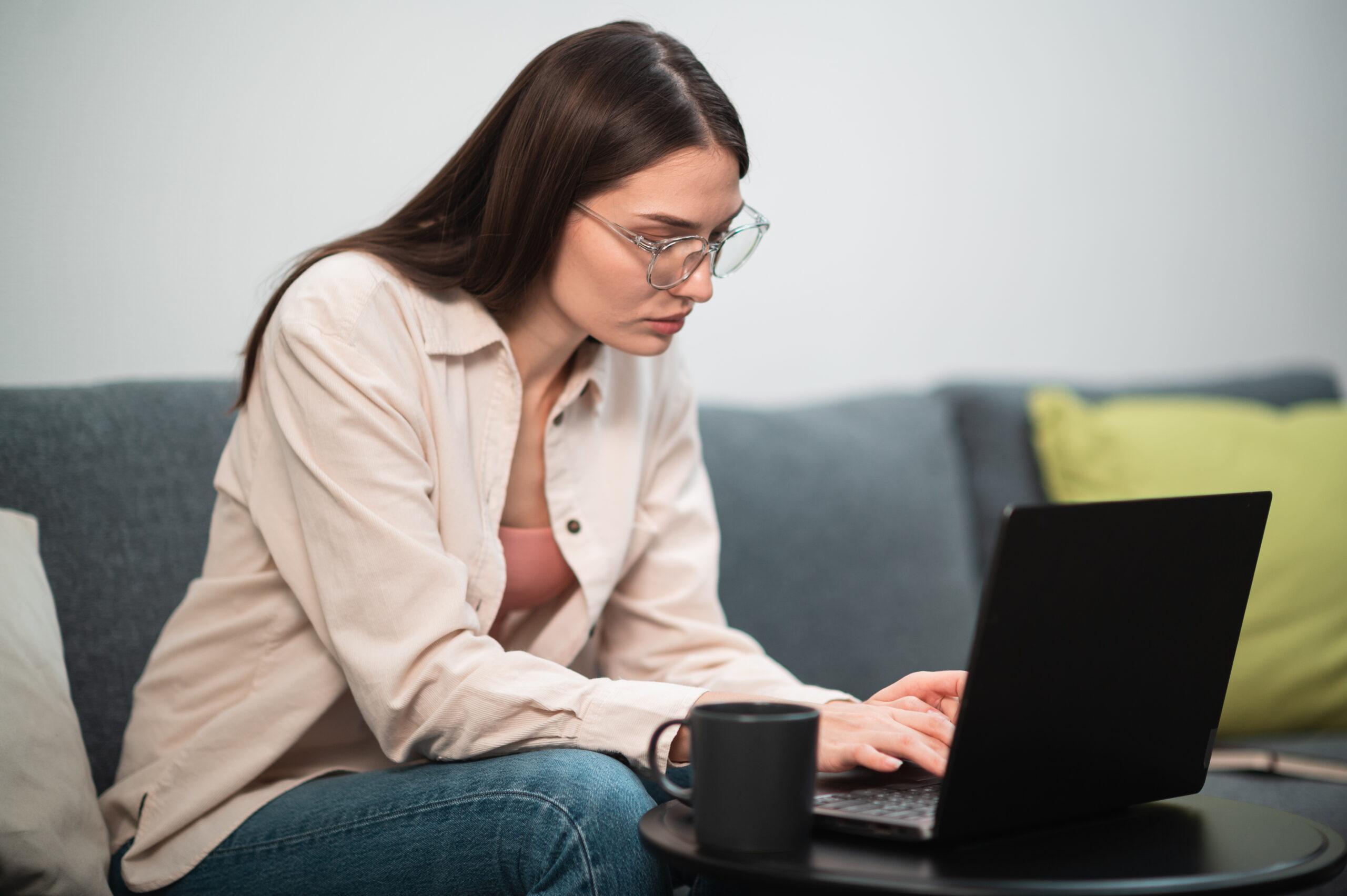 Young woman with glasses sitting on the couch and working on laptop