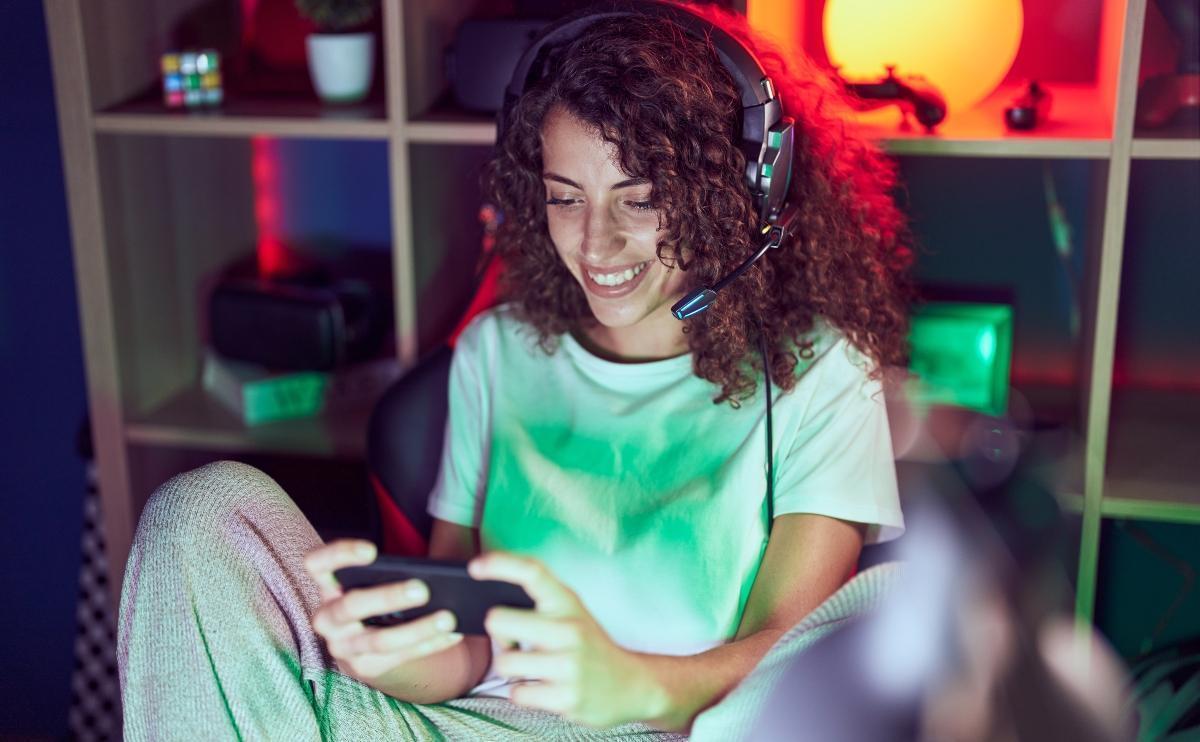 Young beautiful Hispanic woman streamer playing video game using smartphone at gaming room