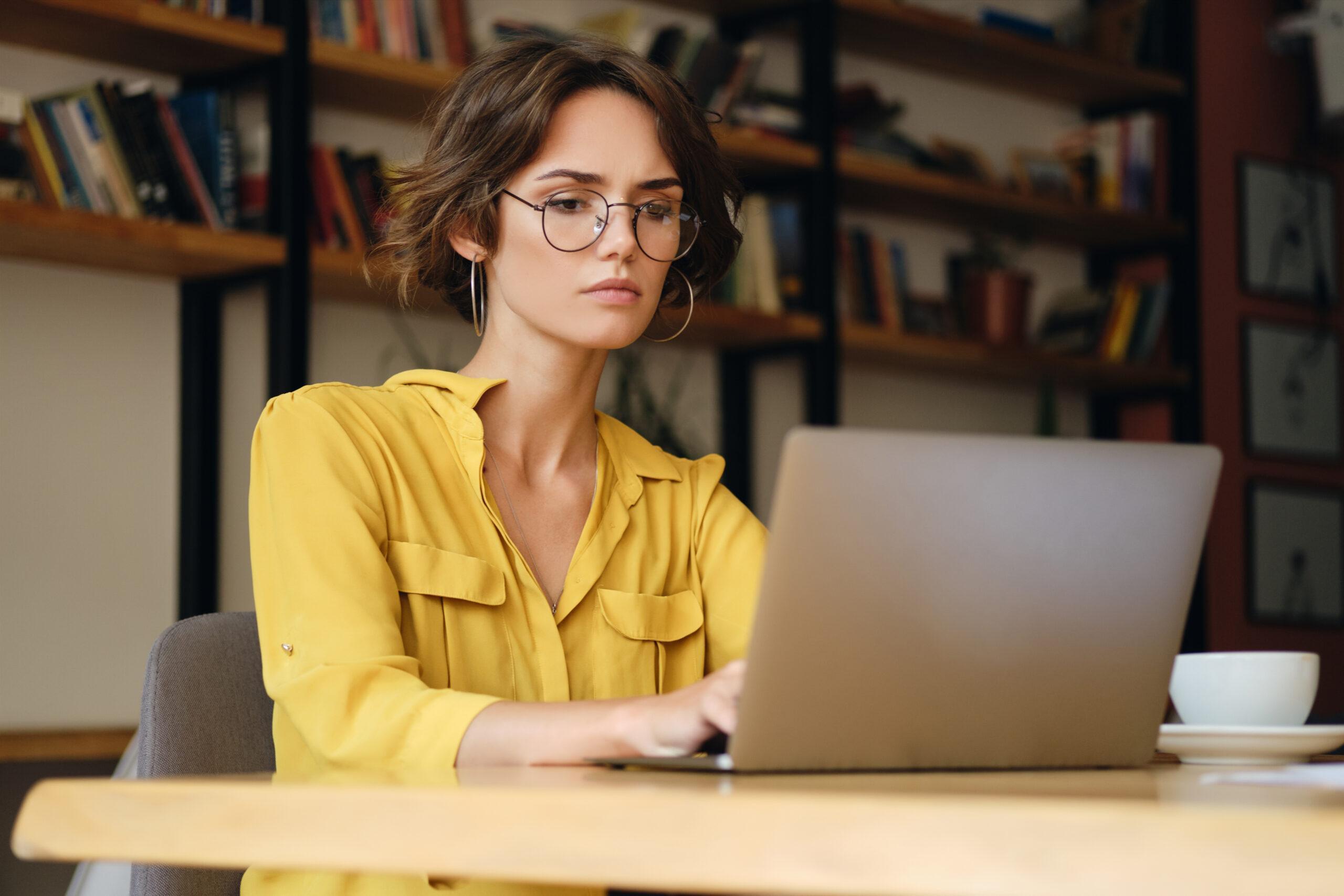 Young serious woman in eyeglasses sitting at the desk thoughtfully crafting an email with laptop in modern office