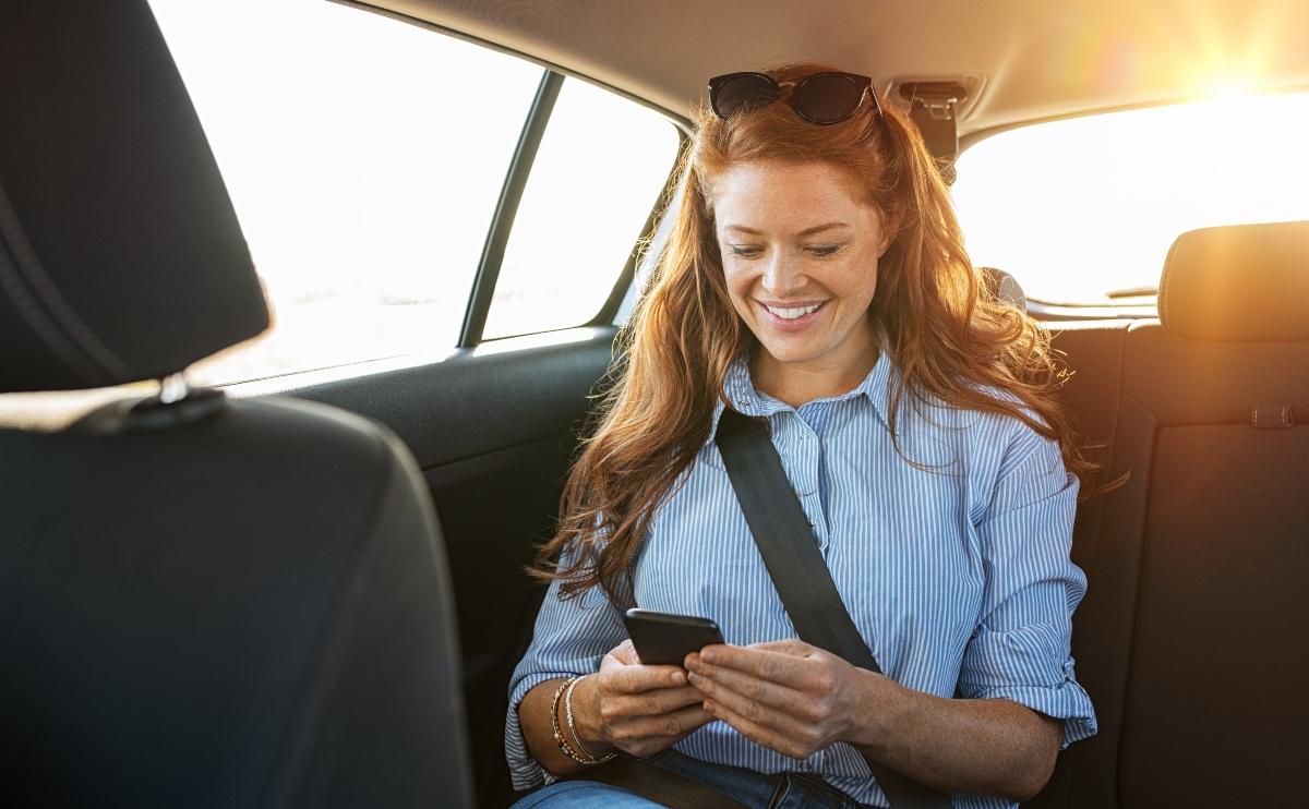 Smiling casual woman sitting in back seat using mobile phone. Cheerful young woman with red hair reading messages in smartphone while sitting at the backseat of a taxi