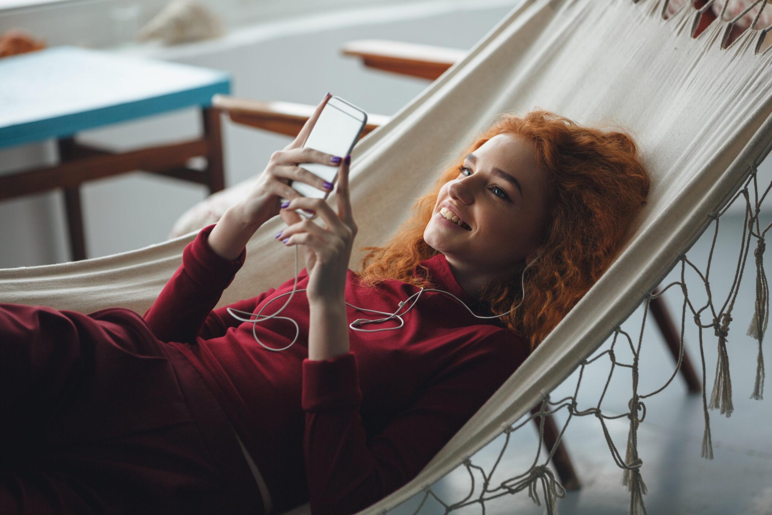 Beautiful happy young redhead lady lies on hammock and chatting with friends on phone