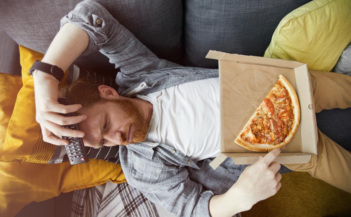 bored man watching tv while lying on a couch and eating pizza