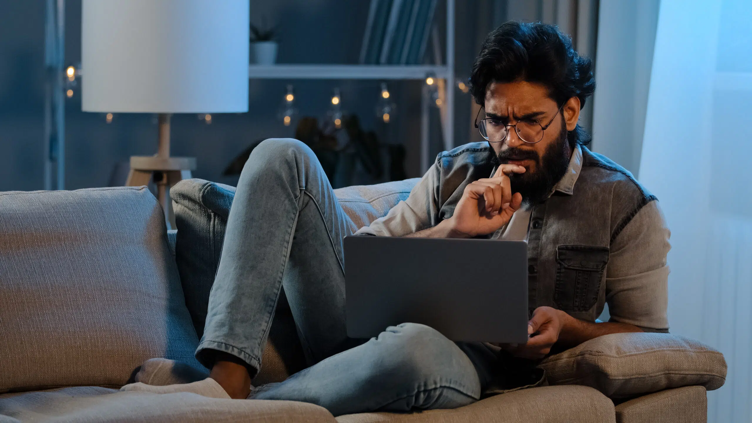 Bearded man wearing glasses sitting on sofa late at night with laptop 