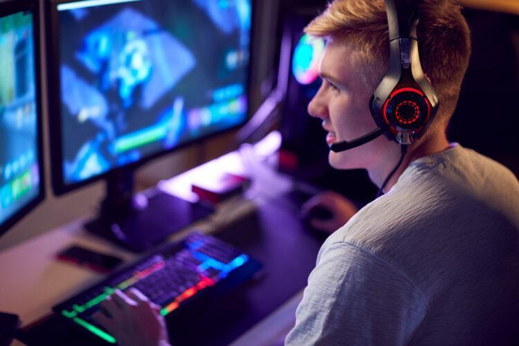 Teenage Boy Wearing Headset Gaming At Home Using Dual Computer Screens