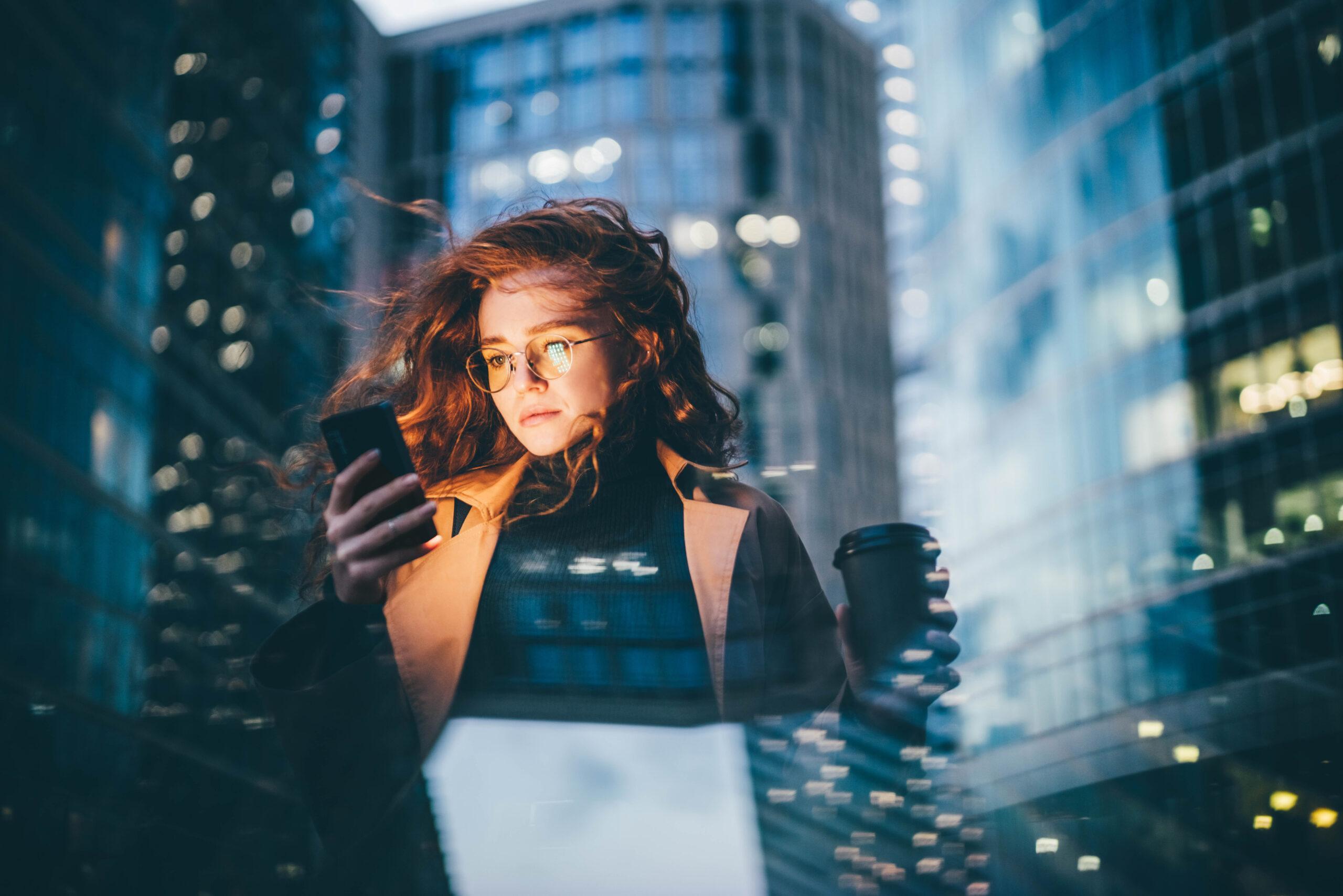 Successful woman using smartphone outdoors while standing near skyscraper at night time