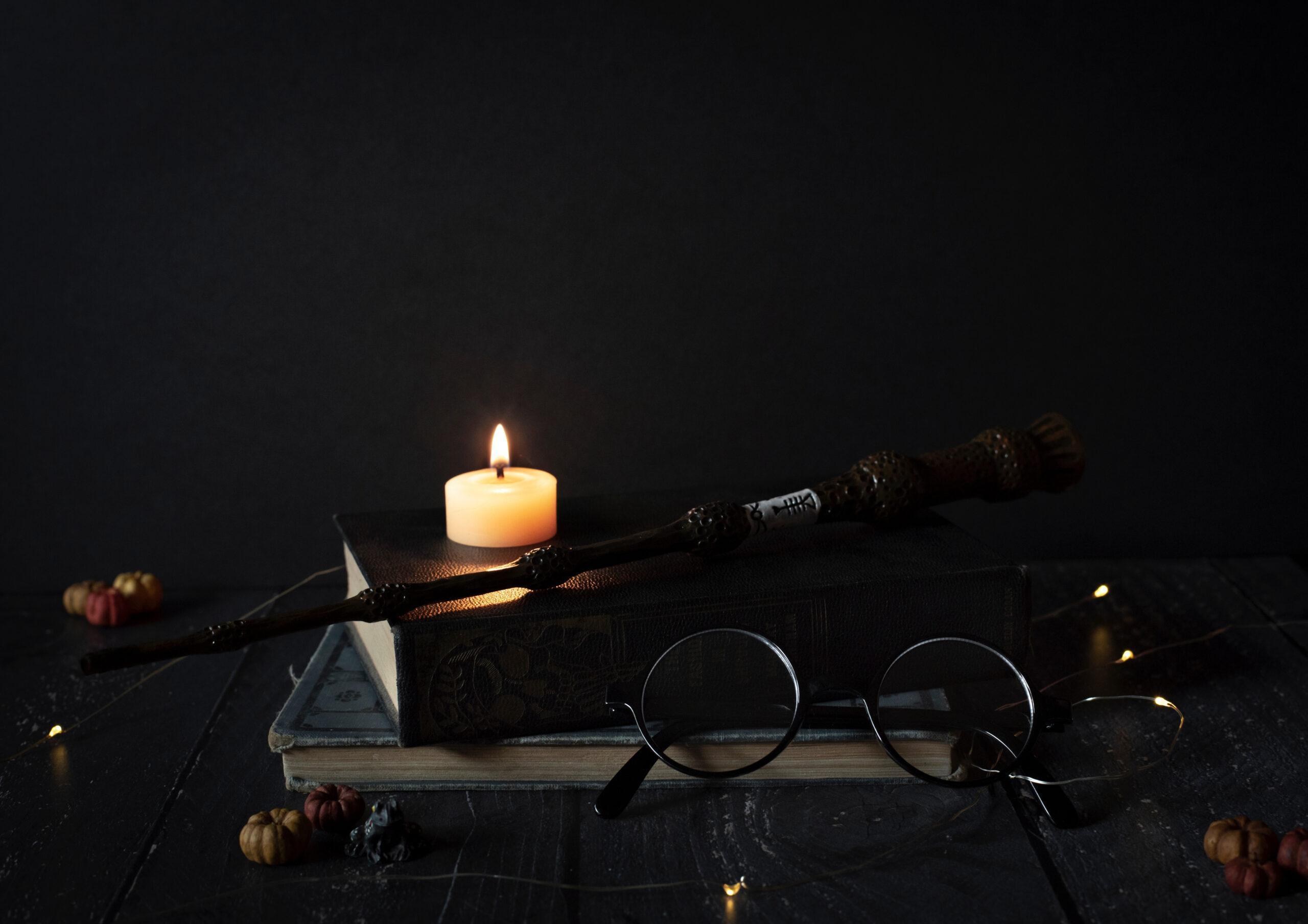 A burning candle, a magic wand on top of books with a pair of glasses 