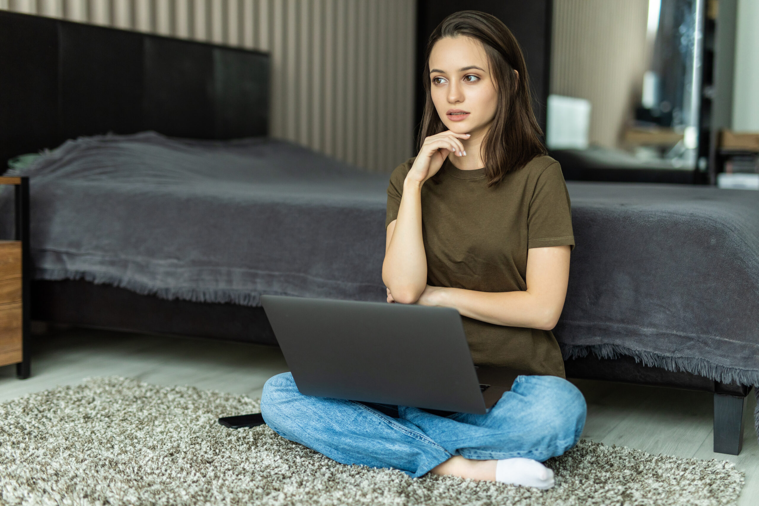 Young woman using computer laptop sitting on the floor with hand on chin
