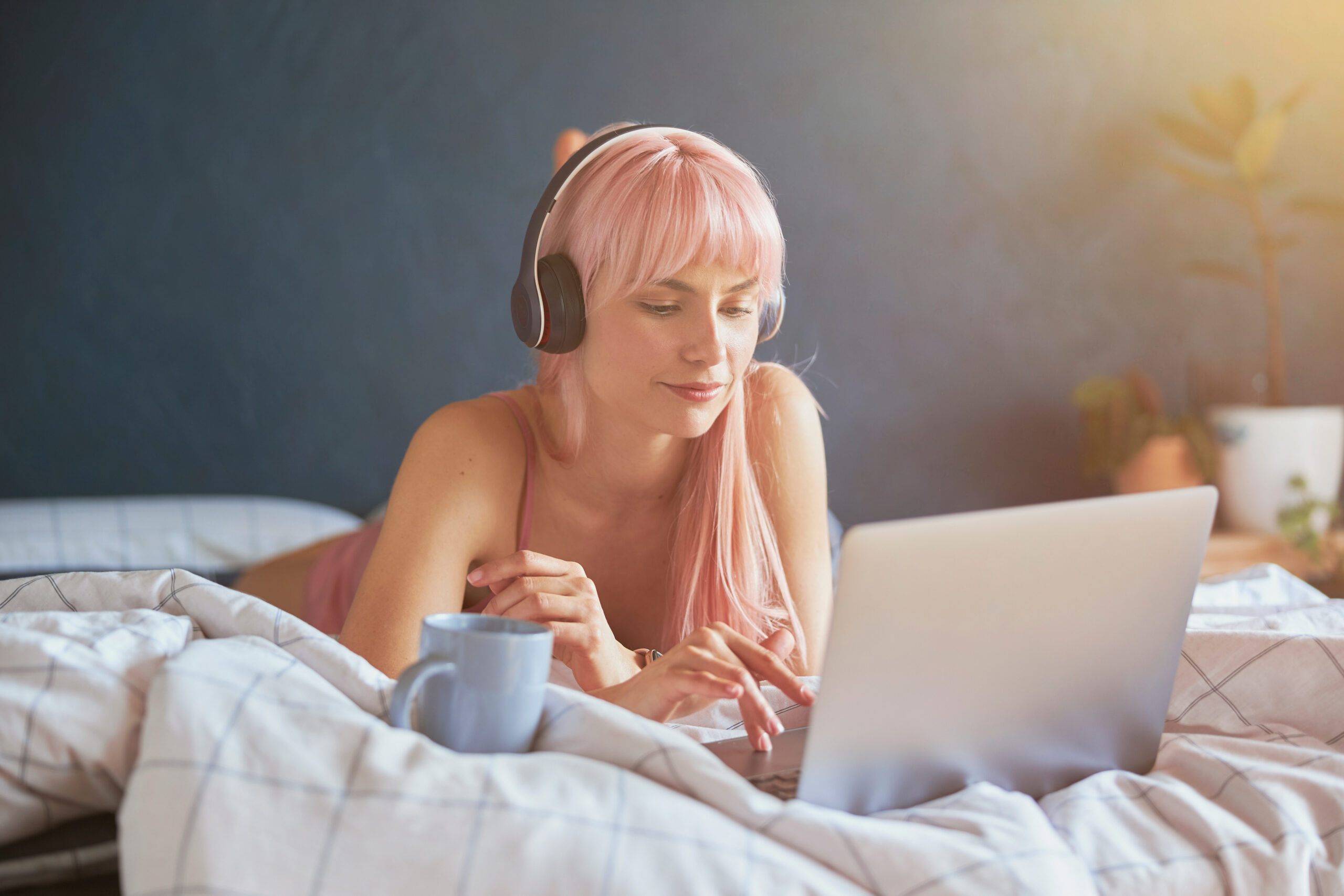 Young female model with headphones and cup works on modern laptop on bed