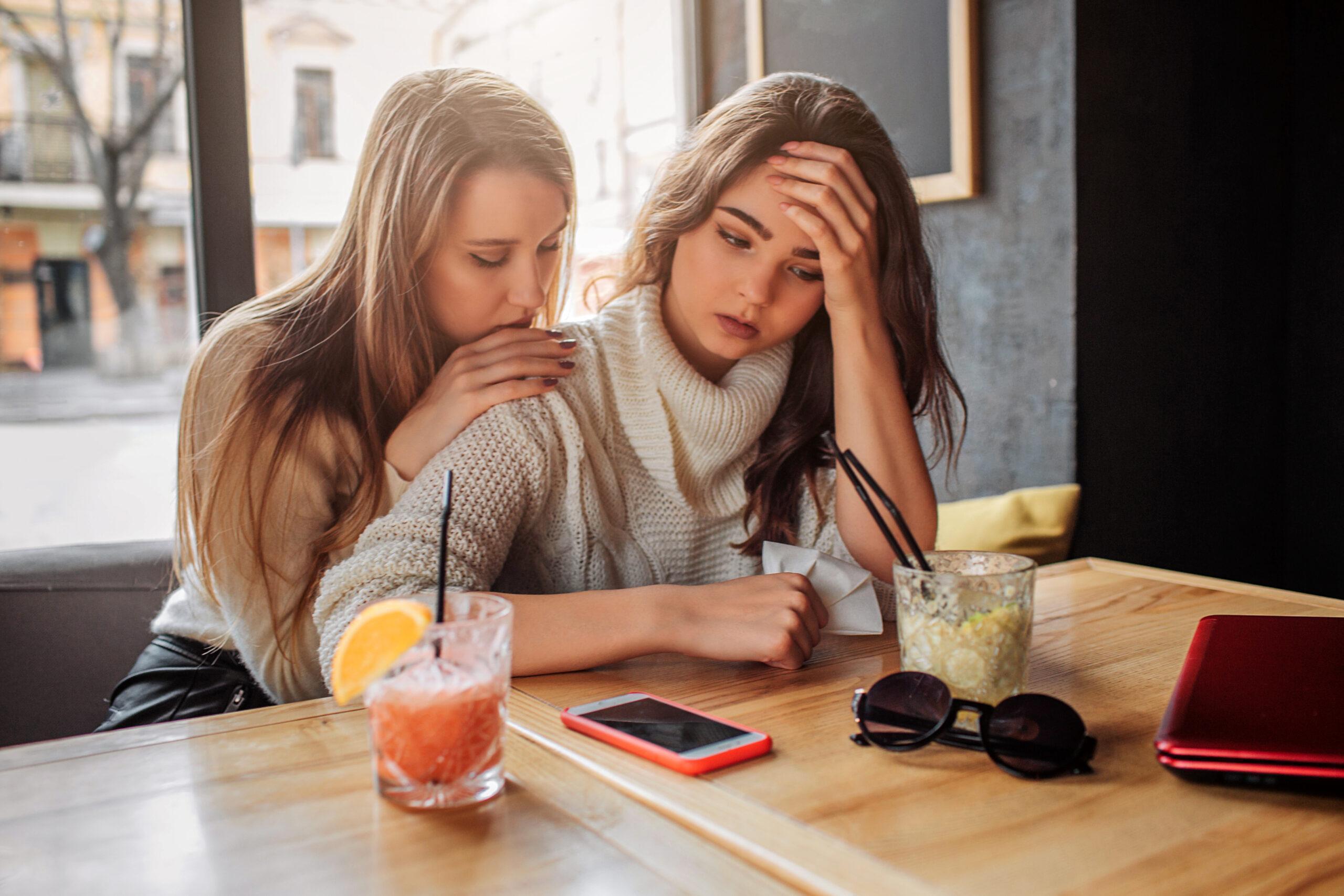 Upset brunette putting hand on head. Blonde woman leaning and trying to comfort her friend.