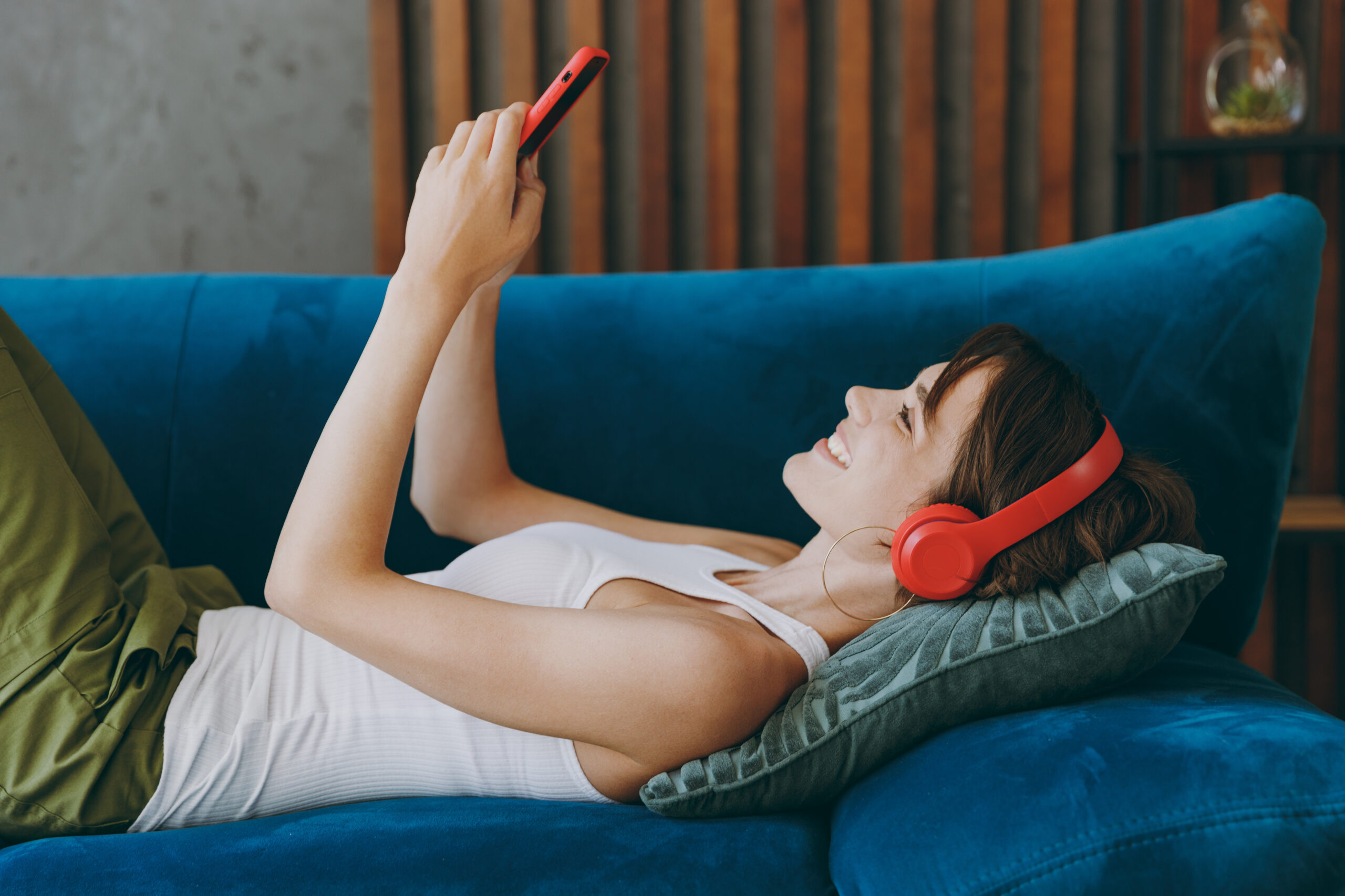 young happy woman with headphones listening to music on cell phone, lying down on the sofa at home