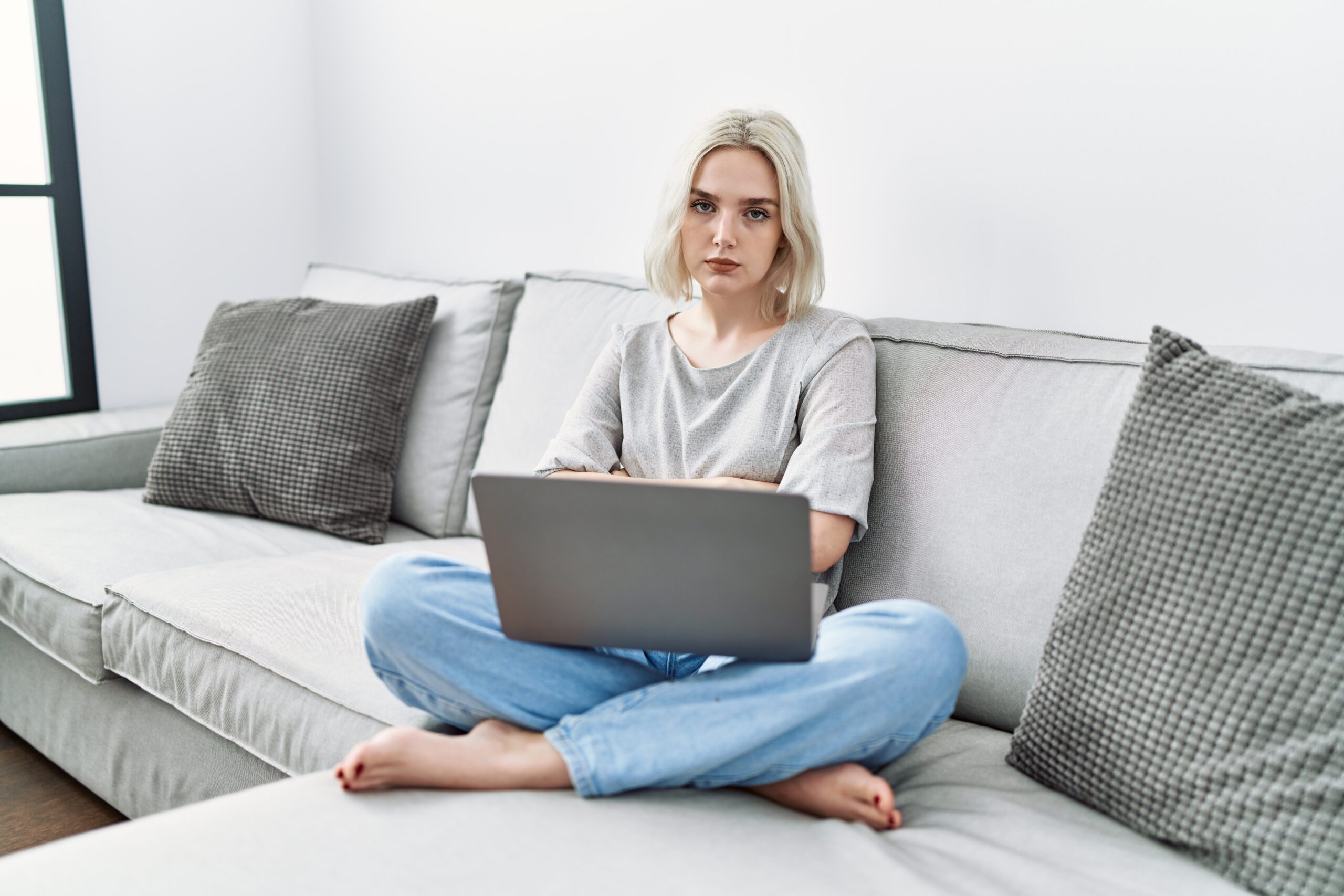 Young Caucasian woman using laptop at home sitting on the sofa with a disapproving expression on her face with crossed arms