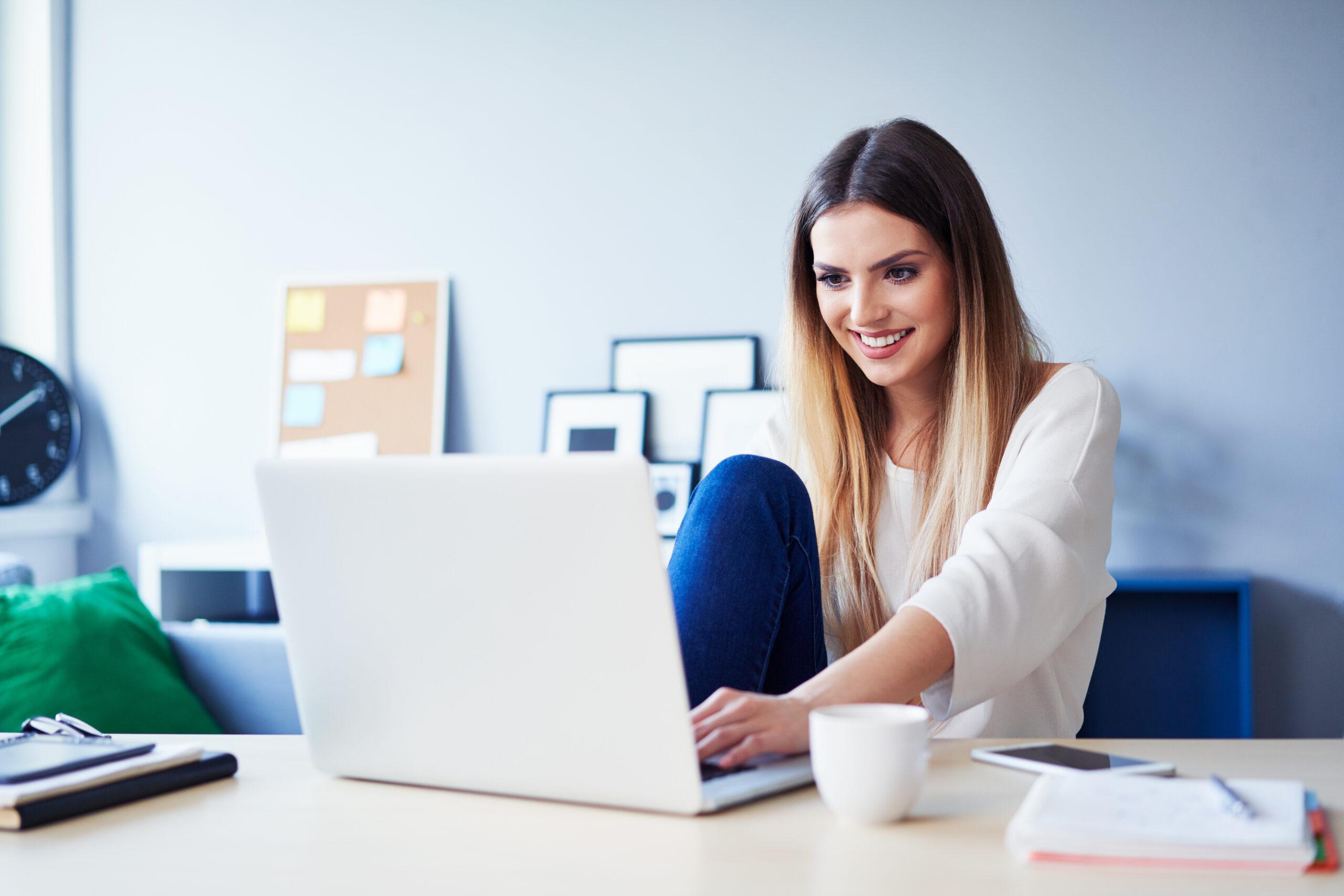 Cheerful young woman using laptop having coffee at desk in home office
