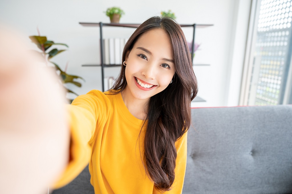 Pretty young asian female with big smile sitting at living room having a video call with someone.