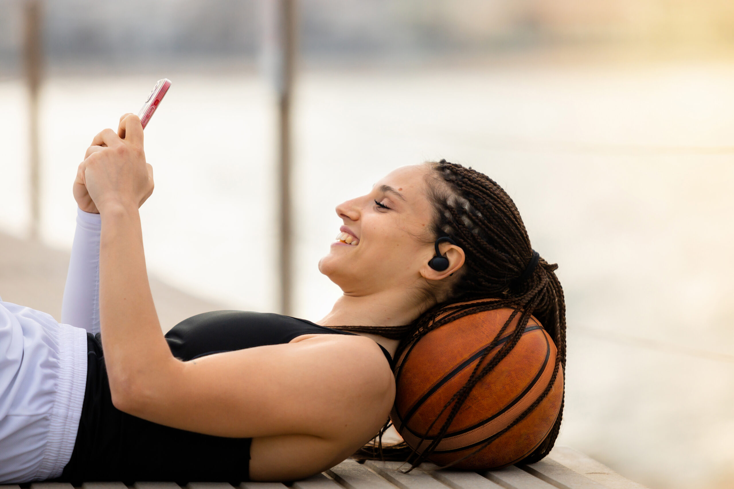 Athletic young woman smiling lying on the ground, head resting on a basketball while playing on her mobile phone