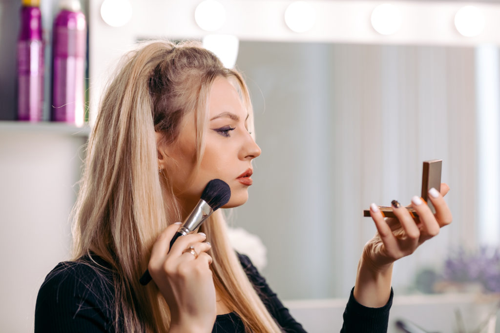 woman applying makeup with powder brush looking in small mirror