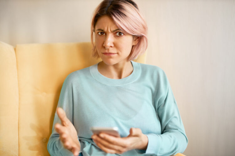 displeased irritated young female in blue sweatshirt sitting on sofa with cell phone, having connection problems or low battery charge