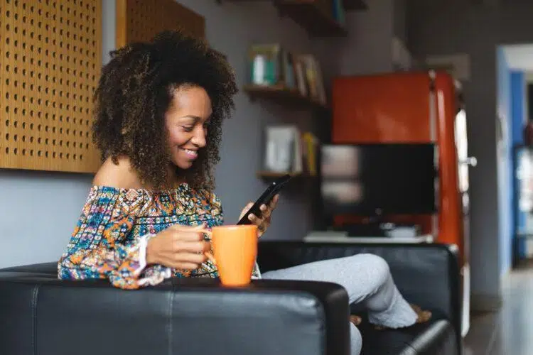 Casual young black woman realxing at home texting on smartphone and drinking coffee