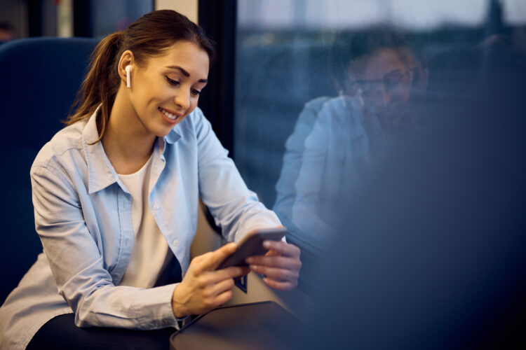 Young happy woman uses cell phone and listens music over earbuds while traveling by train.