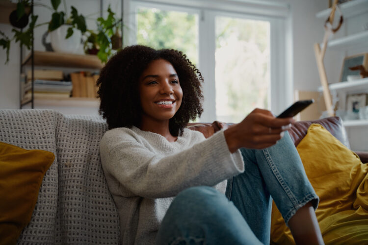 cheerful woman sitting on couch at home watching tv and holding remote control