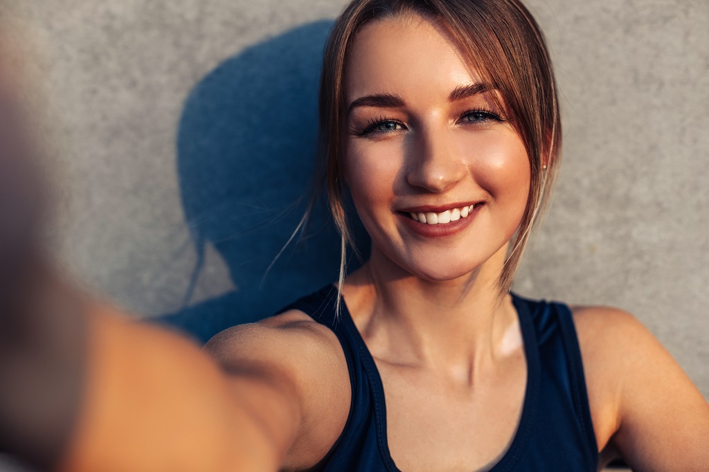 Beautiful athletic woman, taking selfie, photographing herself against gray city wall, sunlight on her face.