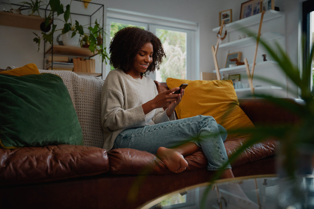 Happy young woman using mobile phone while sitting a couch at home