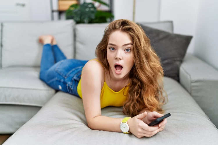Young woman lying on the sofa using smartphone with surprised look
