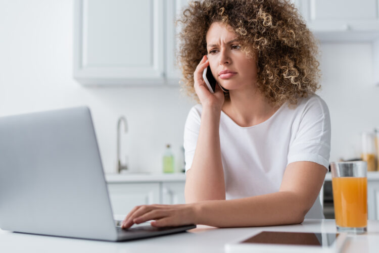 serious and frowning woman using laptop and talking on cellphone in kitchen.