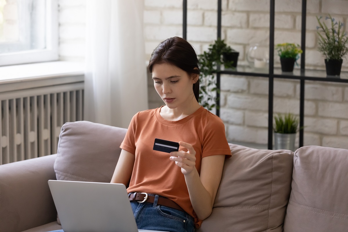 young woman sitting on sofa holding a credit card while looking at the laptop