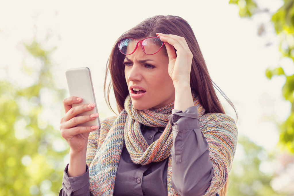Upset woman in glasses looking at her smart phone with frustration while walking on a street