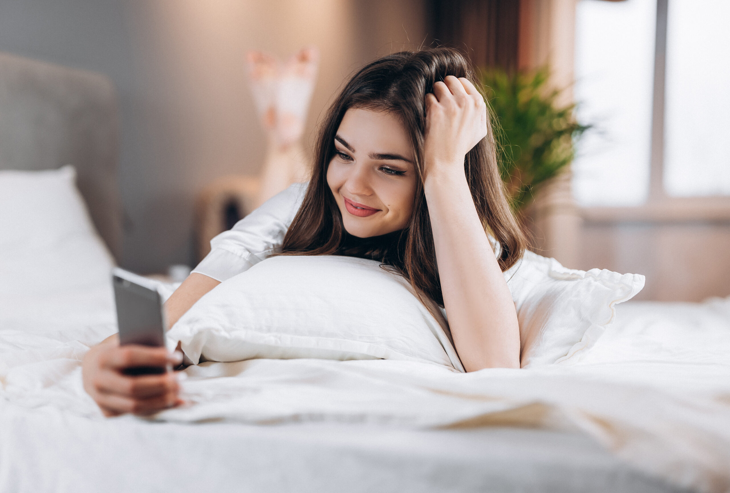 young woman looking at phone lying in bed with a smile on her face