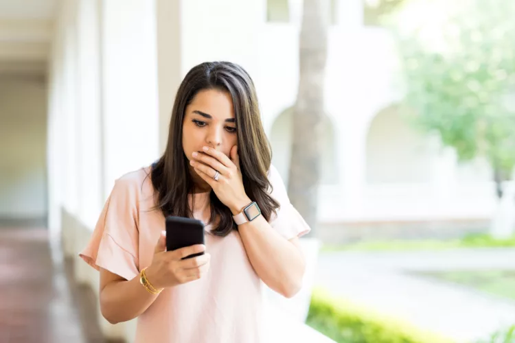 Woman covering mouth while looking at her mobile phone, appears to be surprised

