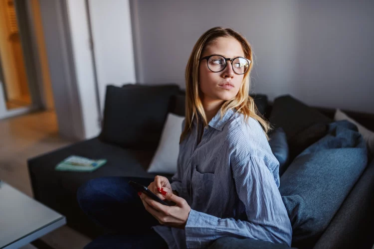 Young serious blond woman with eyeglasses sitting on sofa holding a cellphone