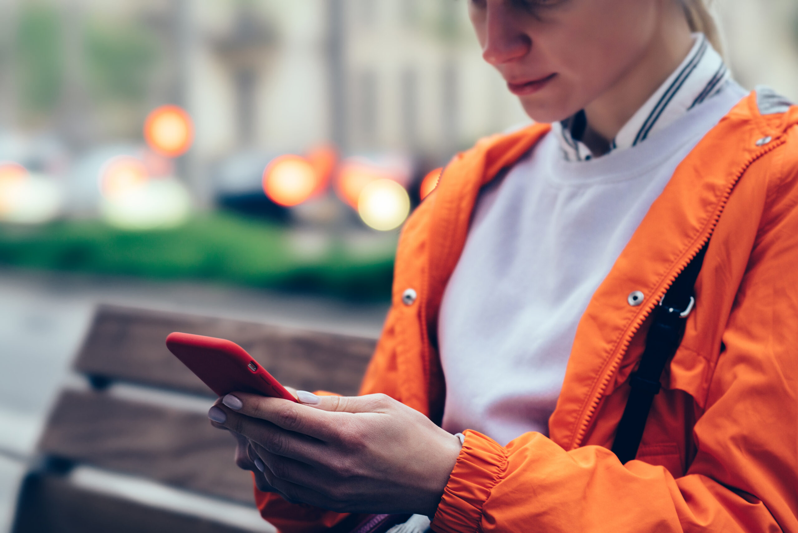 young female holding smartphone in red case while sitting outdoors