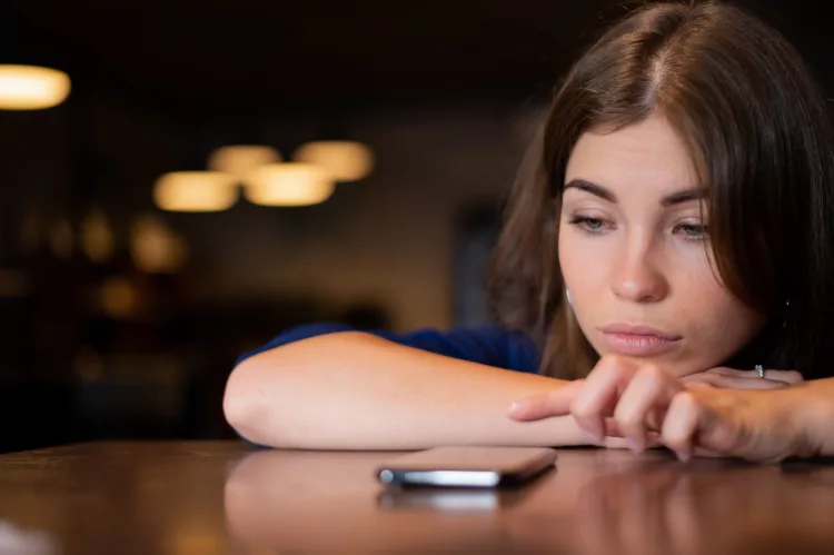 Sad young woman has laid her head on the table and staring at her cell phone on the table
