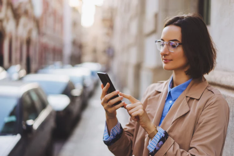Young beautiful woman in glasses and beige coat using mobile phone on the side of the road