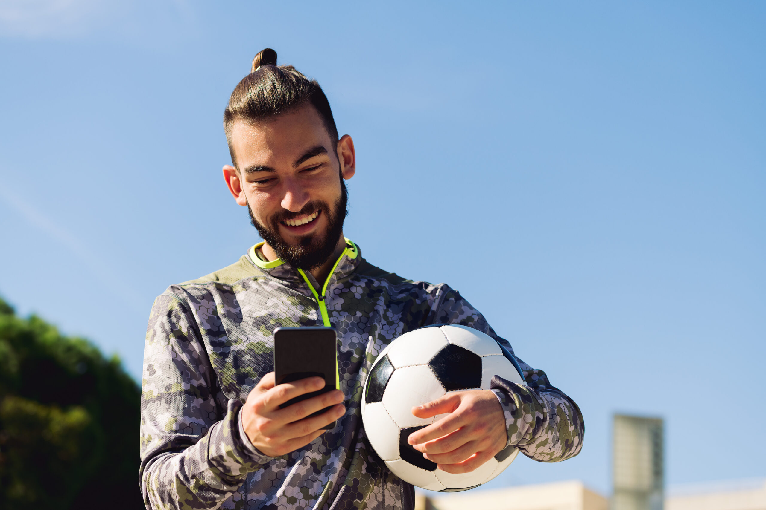 smiling athletic young man holding a football while using his mobile phone