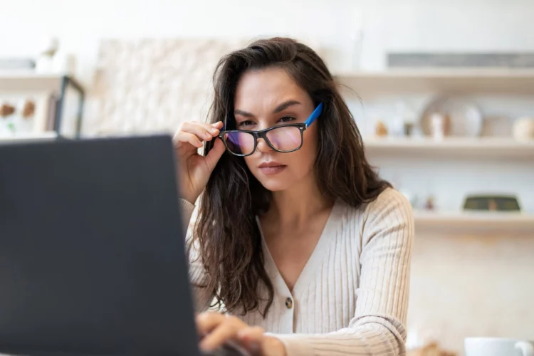 woman lowers down glasses as she looks at laptop screen at kitchen counter.