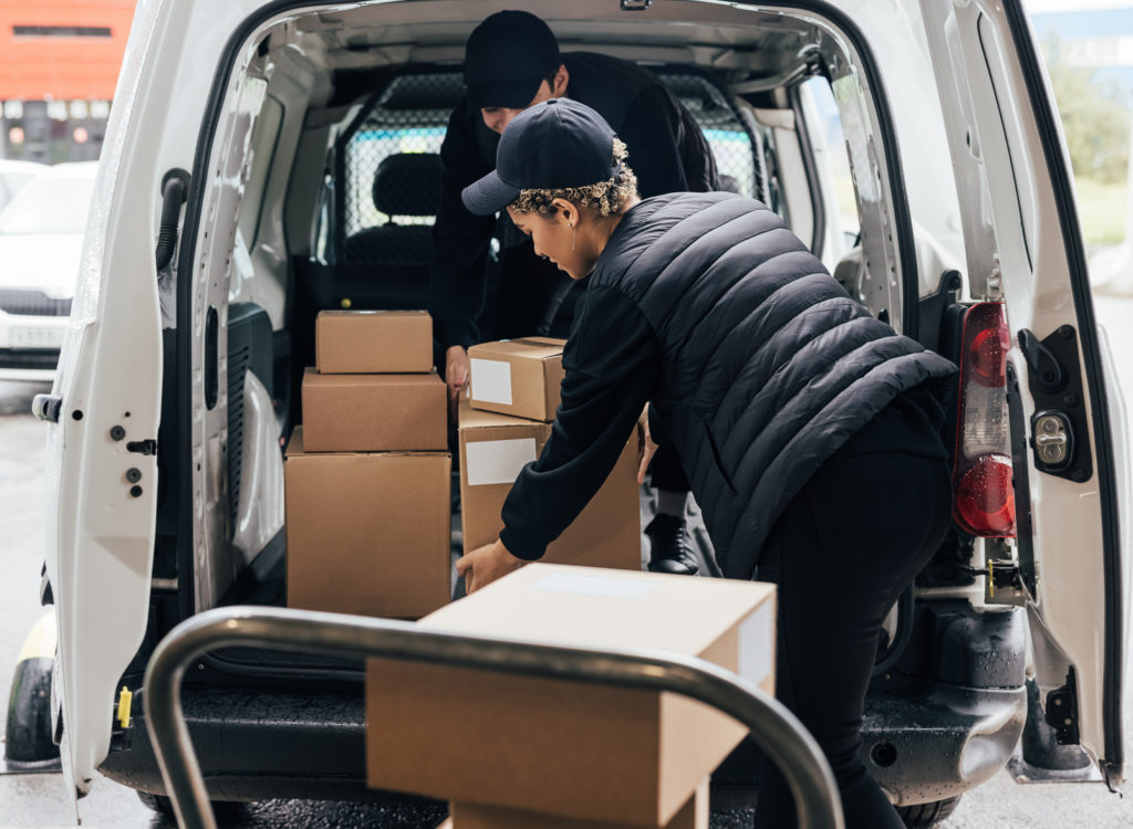 Female courier loads cardboard boxes from a cart into a car working together with a coworker.