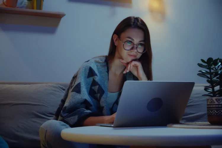Young smiling woman using laptop at night, working overtime at home sitting on sofa.