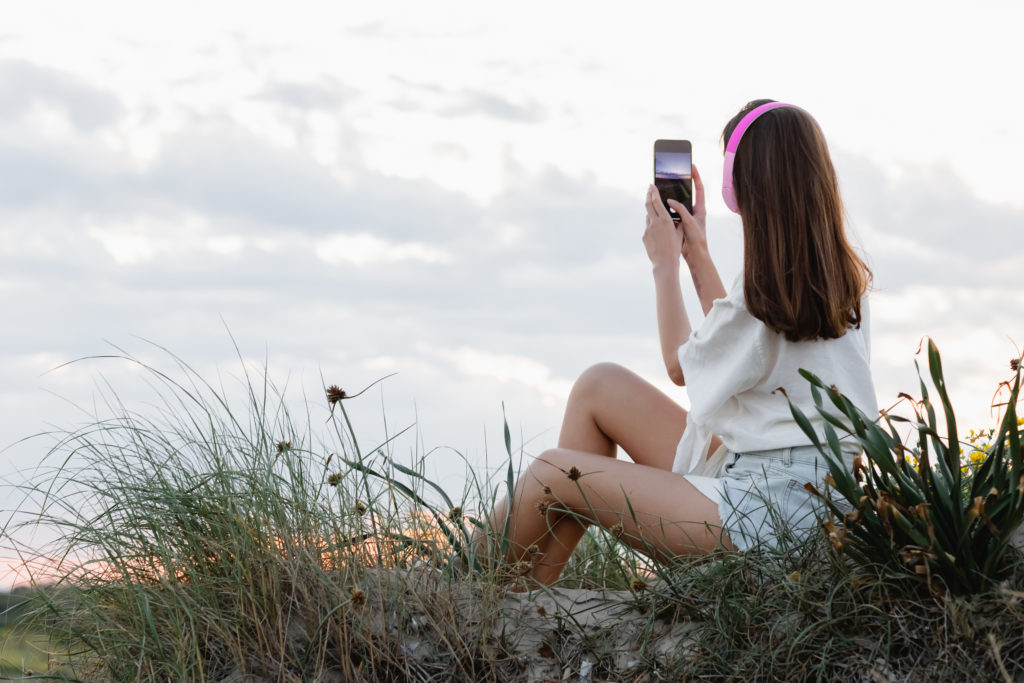 Young woman with headphones seated on the ground taking a photo on smartphone outdoor.