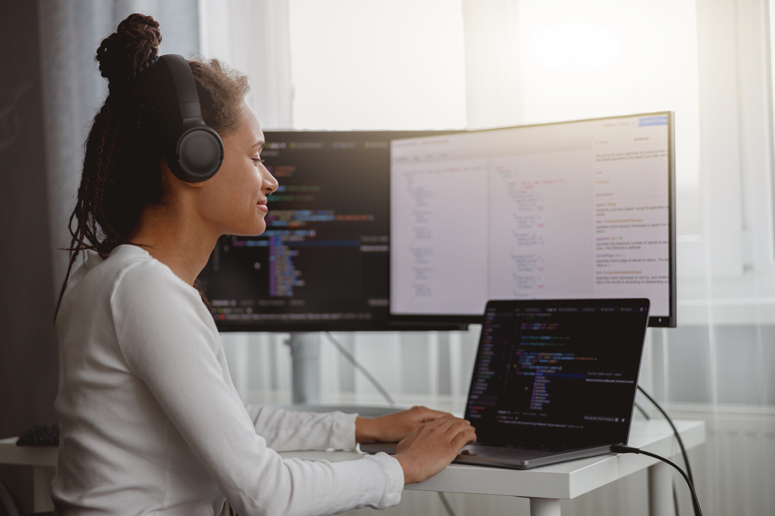 Cheerful beautiful female IT developer listening to music in headphones while coding at home