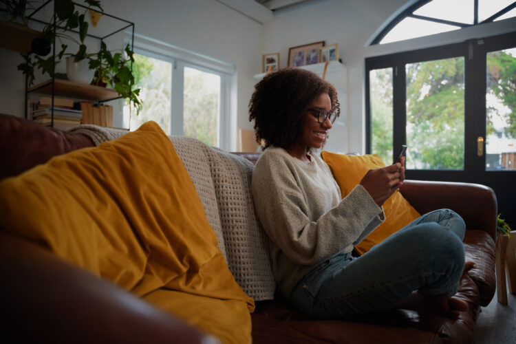 Young african woman wearing spectacles using smartphone at home sitting comfortable