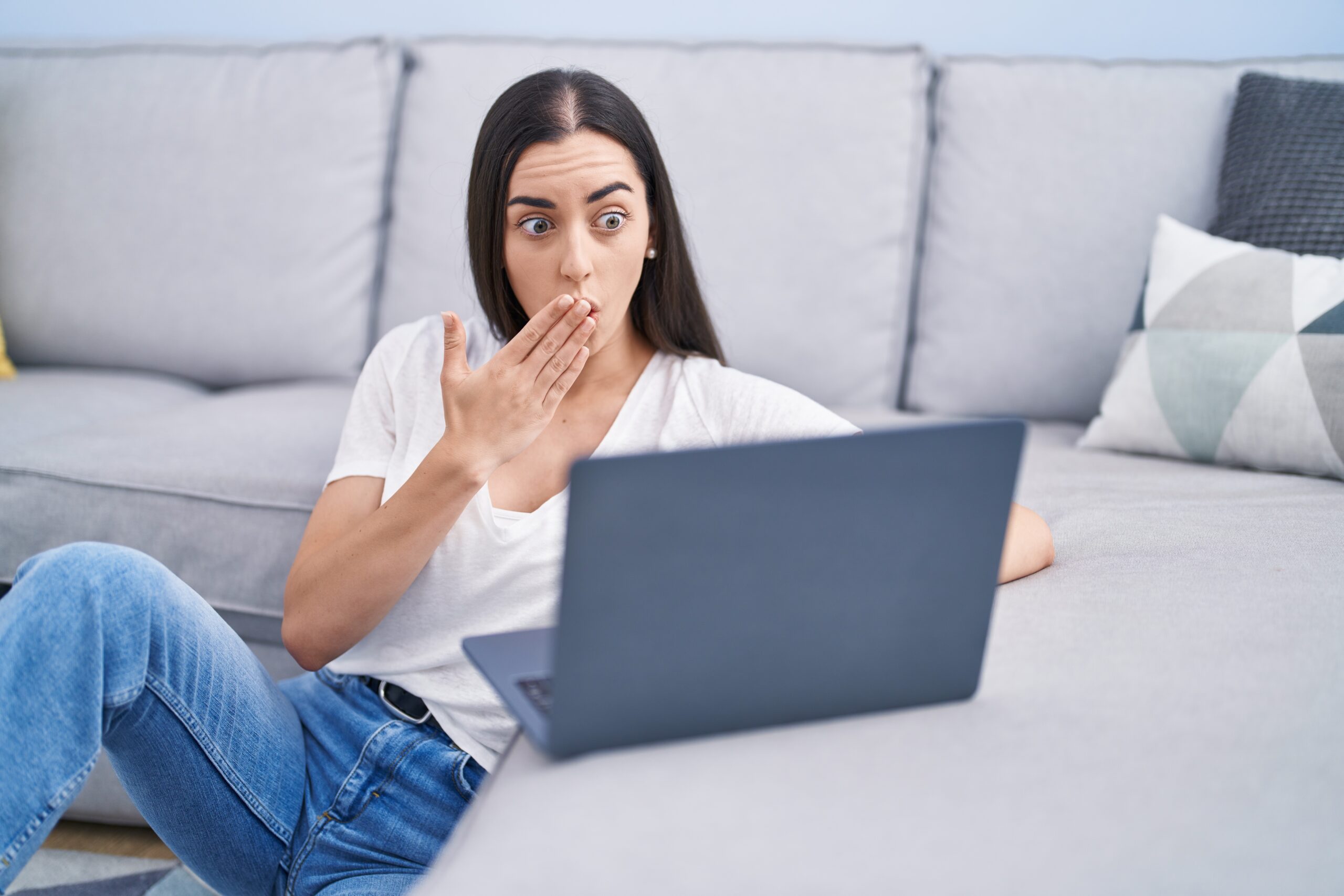 Young brunette woman using laptop at home covering mouth with hand, shocked and afraid for mistake