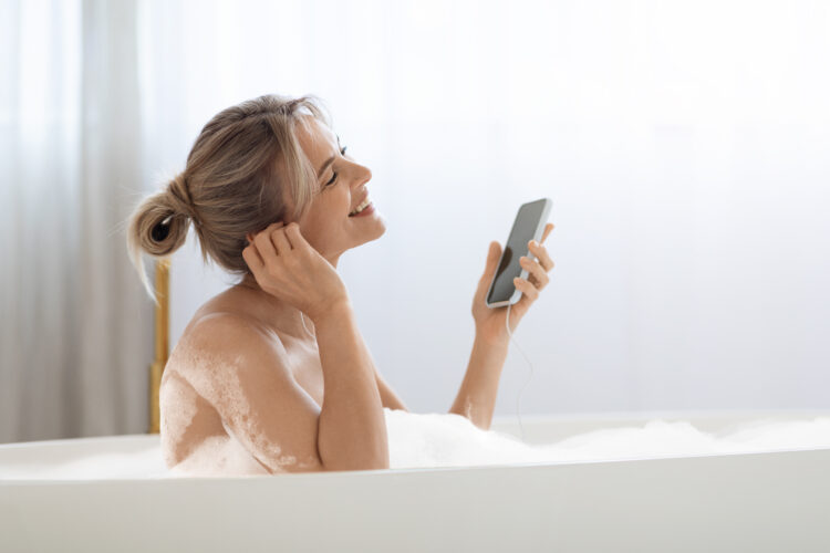 Happy blonde woman taking foamy bath and listening to music on the phone.